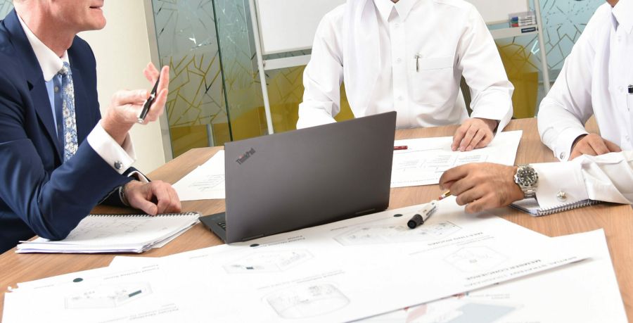 Close up of three people sitting a table, with many papers and a computer, having a discussion