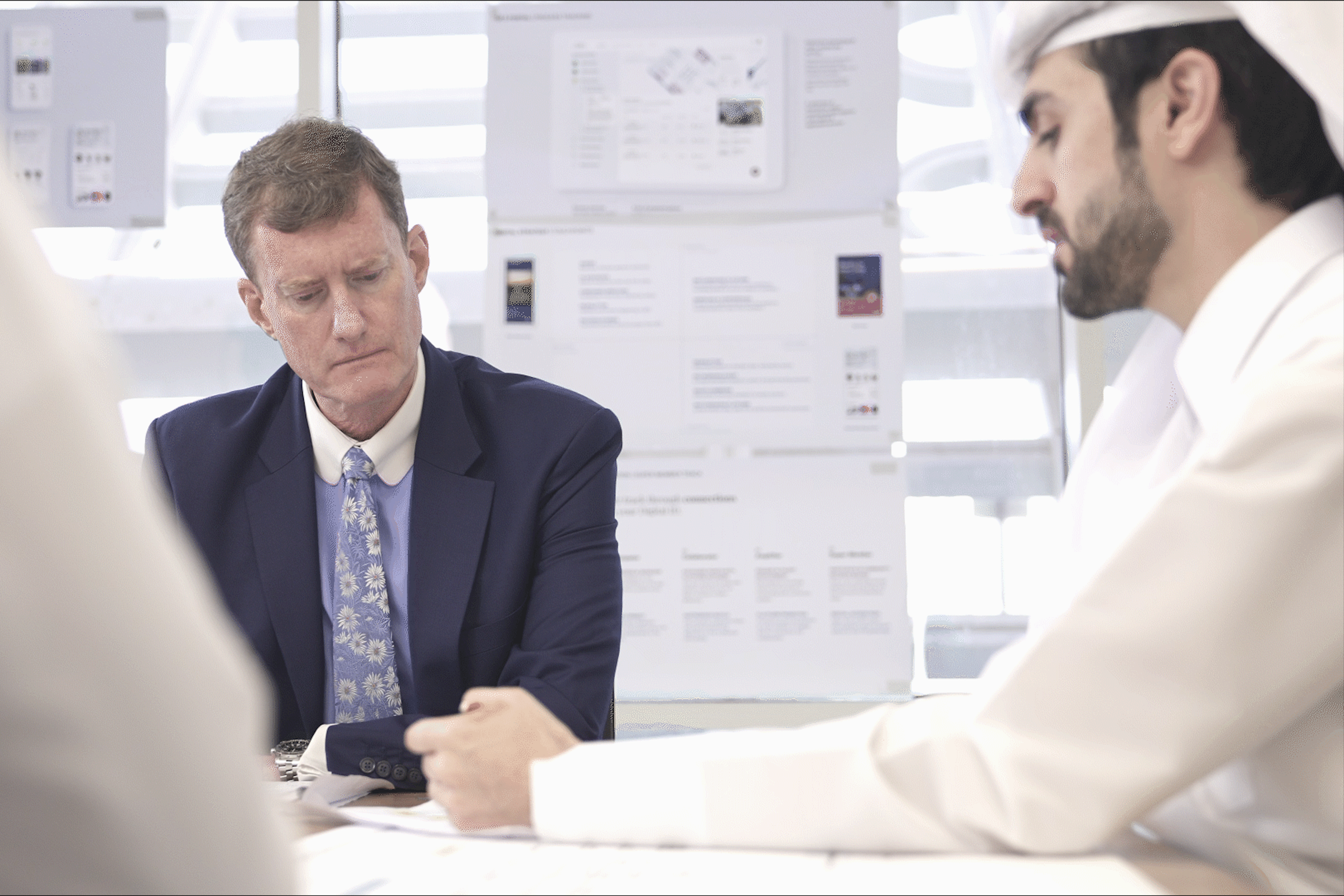 Two businessmen, one in a suit and one in traditional Arabic clothing, sitting at a table in a conference room full of documents, discussing QFZ investment processes