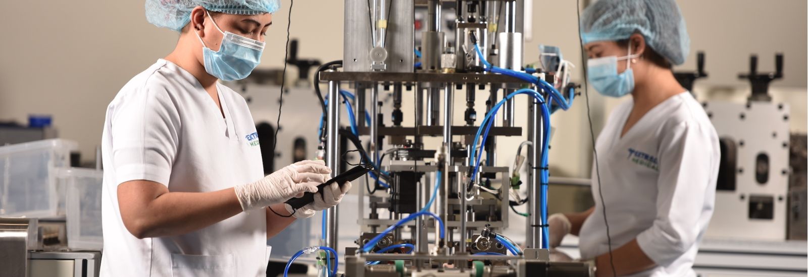 Two people, in white uniforms with hairnets and masks, working on testing manufacturing equipment